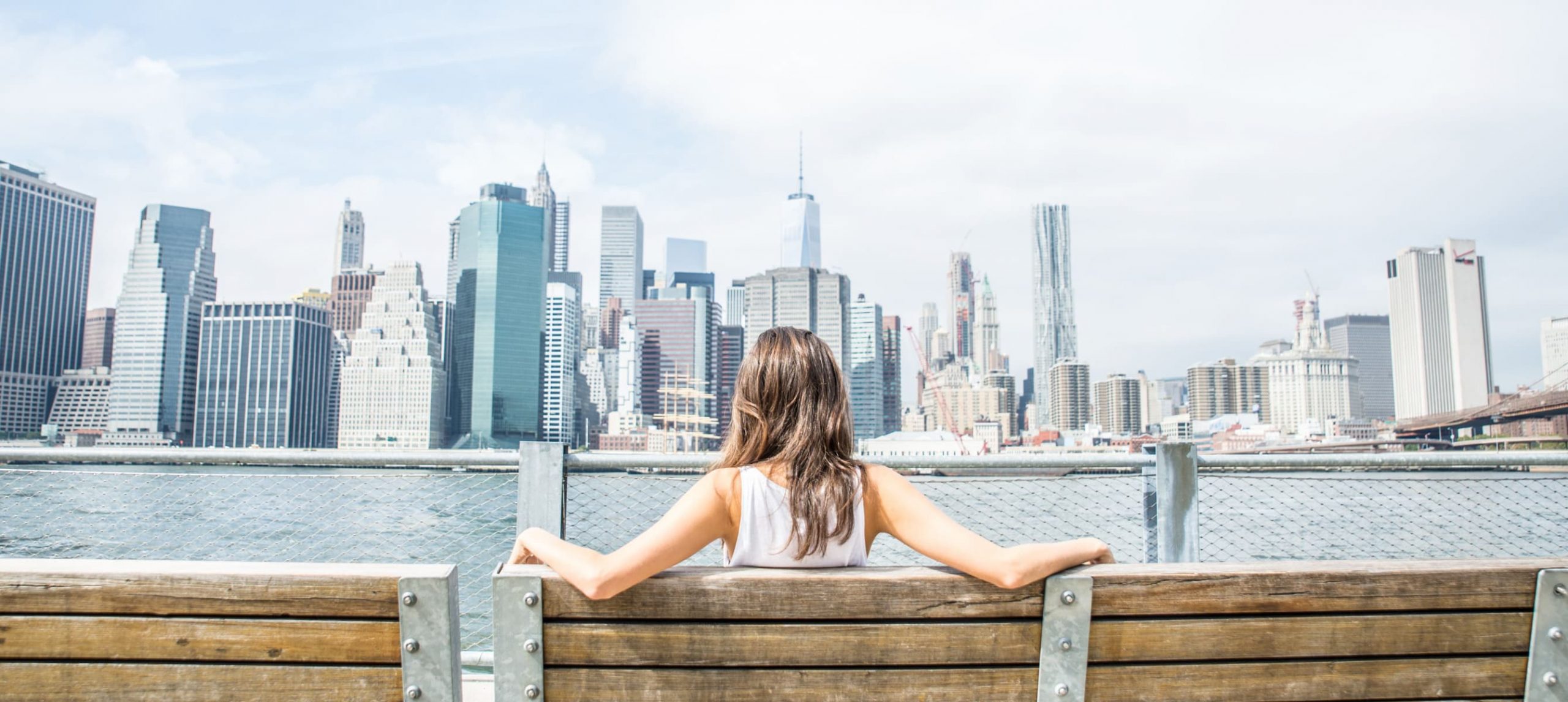 Young woman sitting on a bench and looking at New York skyline.