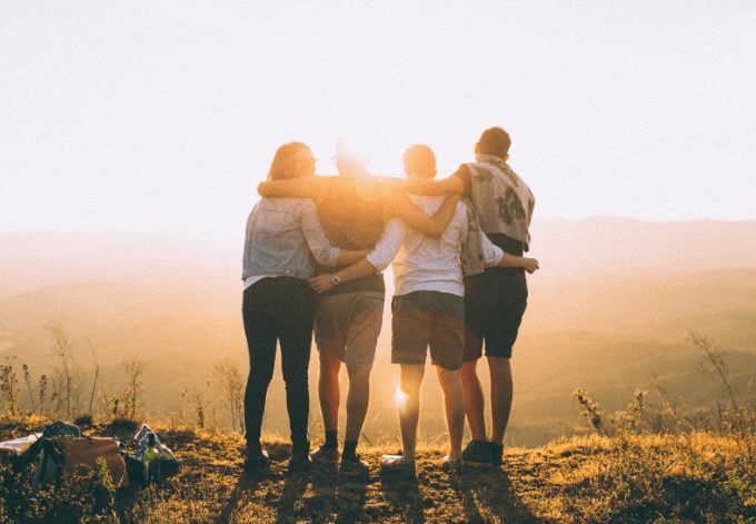 Four friends hugging each other and overlooking a natural landscape at sunset.