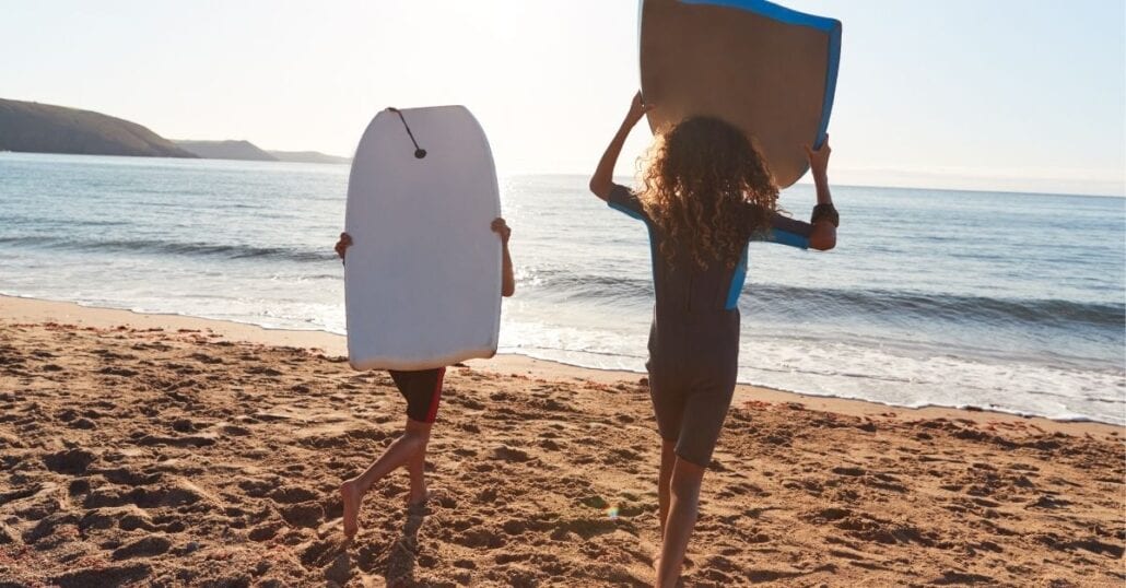 Two kids playing with bodyboards on a sandy beach during a clear afternoon.