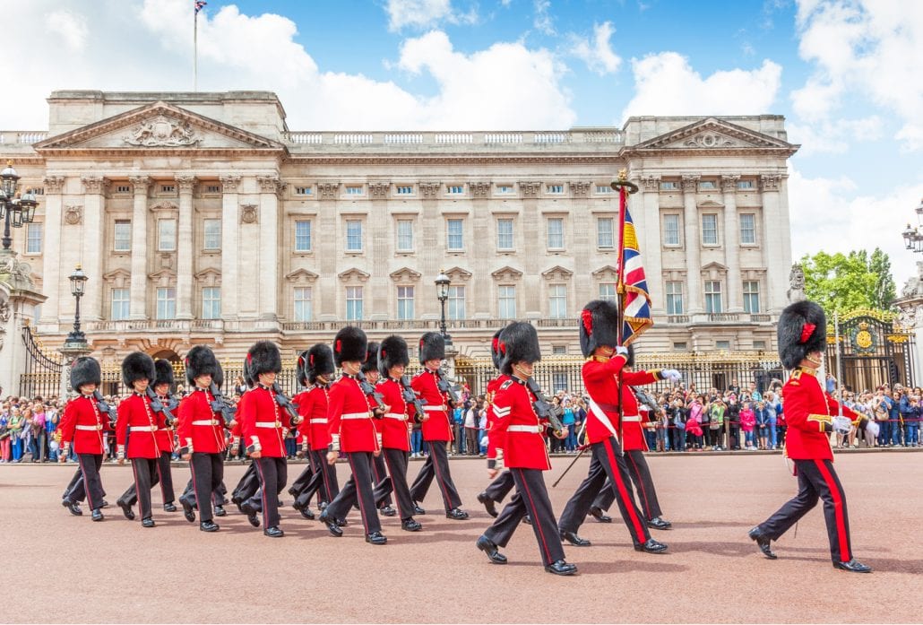 Some 20 guards marching in front of the Buckingham Palace while being watched by a crowd of people.