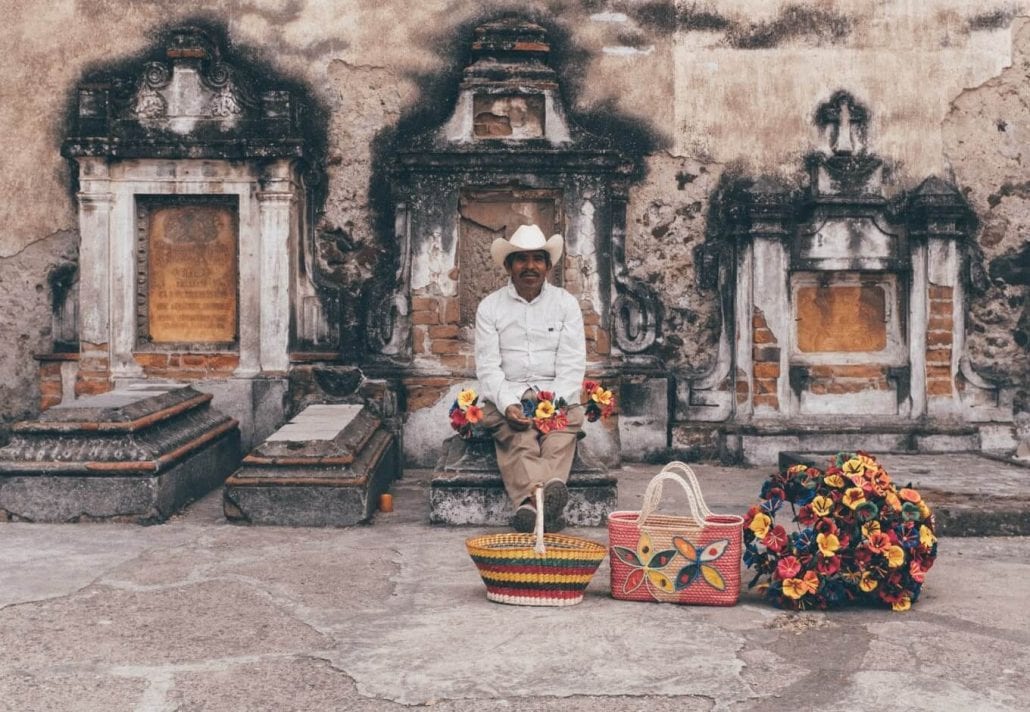 Portrait of a Mexican man peacefully sitted on a grave, in Mexico, and holding colorful flowers. 
