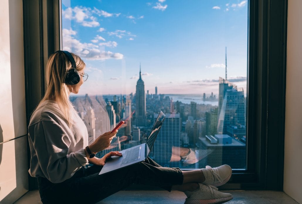 Blond woman with headphones on, a laptop on her lap, sitting on the windowsill and looking at New York's skyline.