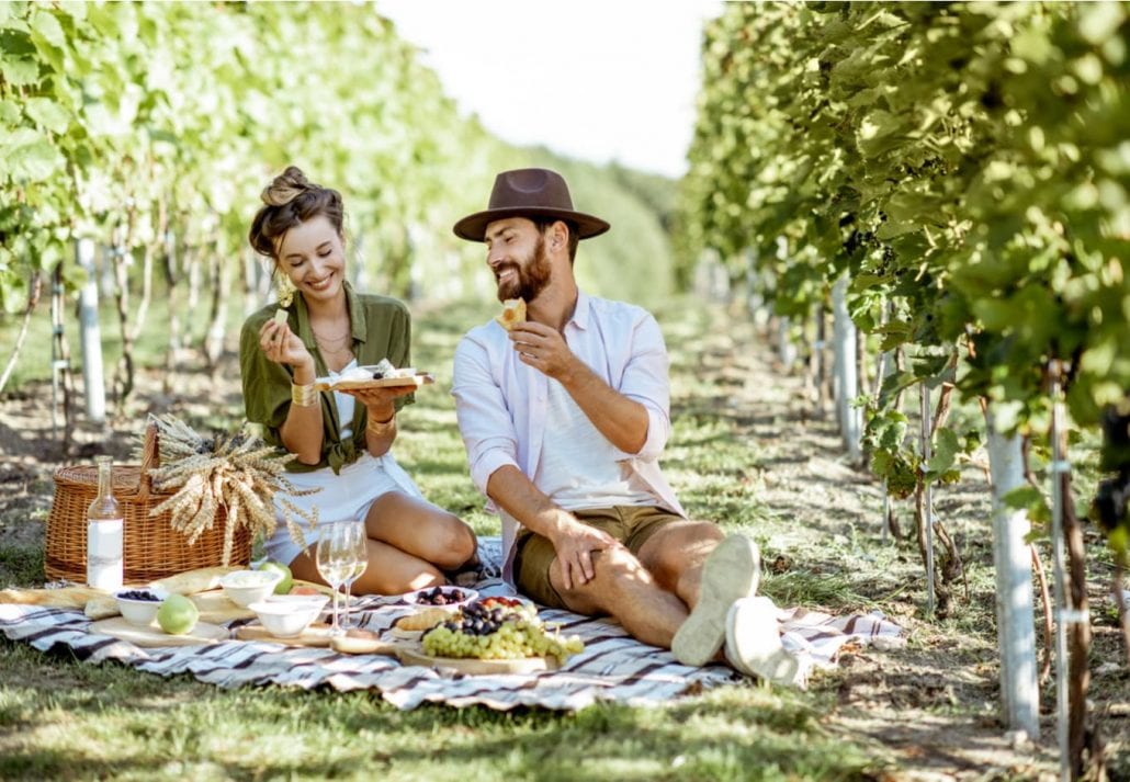 Beautiful couple having romantic breakfast with lots of tasty food and wine, sitting together on the picnic blanket at the vineyard on a sunny morning
