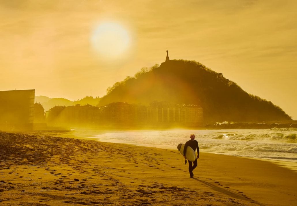 A surfer walking on the Zurriola Beach at sunset with the Monte Urgull in the background. San Sebastian, Basque Country, Guipuzcoa. 