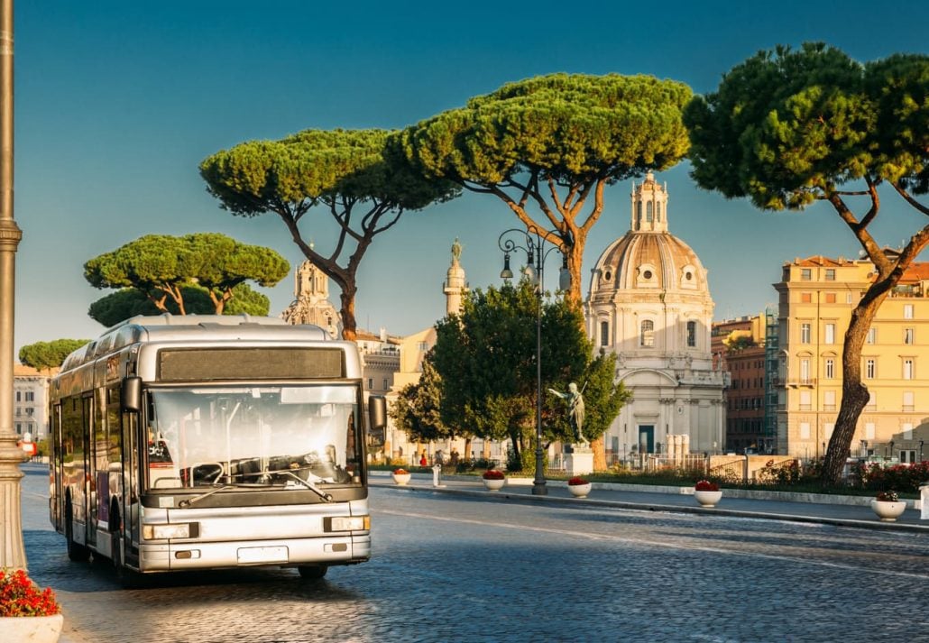 Public City Bus At Bus Stop In Via Dei Fori Imperiali Street In Sunny Summer Morning. View On Church Of Most Holy Name Of Mary.
