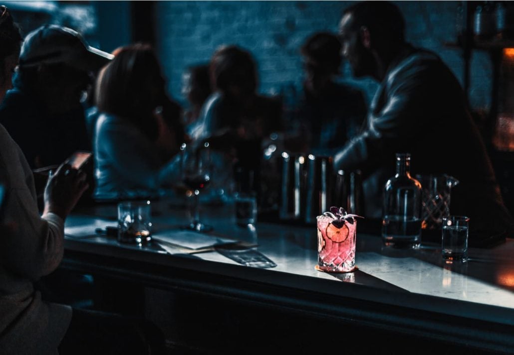 People chatting at the table in a dark bar with a pink cocktail in front of them