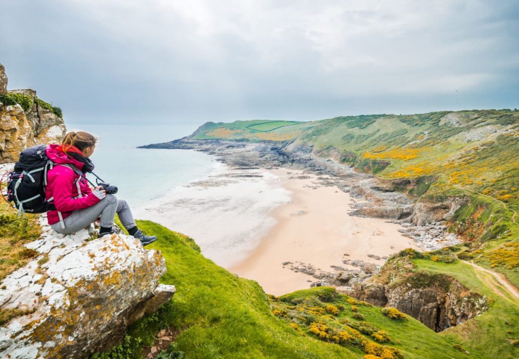 Female Backpacker Looking Into Distance From A Cliff At Rocky Sea Shore On A Cloudy Day In Rhossili, Wales Coast Path, Wales, UK.